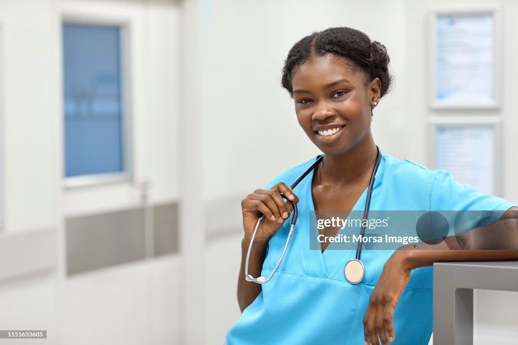 Portrait of smiling female doctor wearing blue scrubs. Young professional with stethoscope is standing in hospital. She is with confident look on her face.